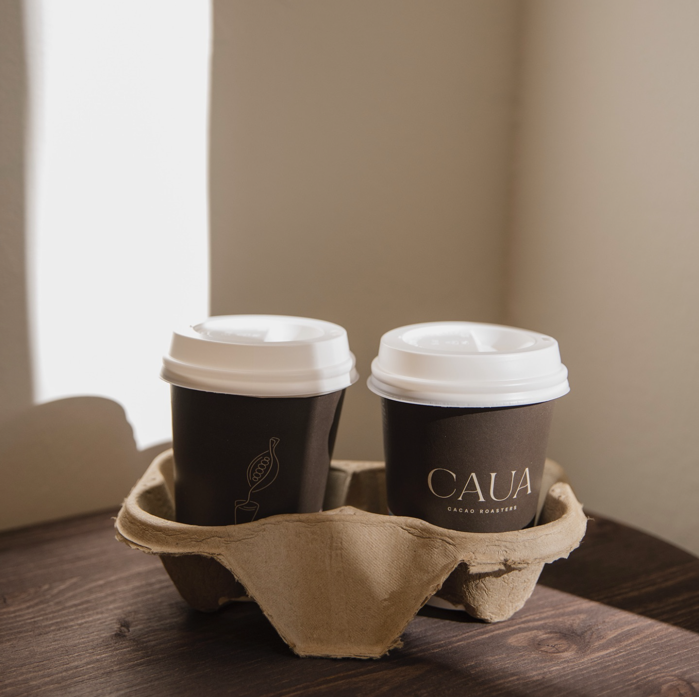 Two dark takeaway coffee cups with white lids, placed in a cardboard cup holder, featuring minimalist branding for a café.